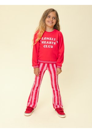Jong meisje met lang blond haar draagt een rood sweatshirt met de tekst "Lonely Hearts Club", roze en rode gestreepte broek en rode sneakers, glimlachend.