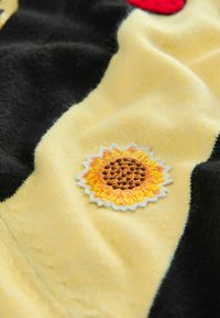 Yellow fabric with a soft texture features a sunflower embroidery patch in orange and brown, surrounded by black and red contrasting areas.