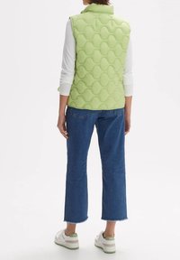 Jasnozielona pikowana kamizelka z wysokim kołnierzem, w połączeniu z białą koszulą z długim rękawem i niebieskimi skrojonymi dżinsami z surowym wykończeniem oraz casualowymi sneakersami.