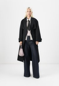Giovane donna con lunghi capelli biondi che indossa un trench coat nero, una camicia bianca, una cravatta nera e jeans a gamba larga, mentre tiene una piccola borsa rosa.