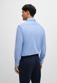 Light blue long-sleeve shirt with a classic collar, smooth texture, buttoned cuffs, and a straight back. Paired with dark trousers.