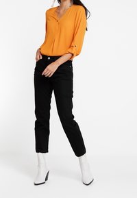 Orange blouse with a V-neck and three-quarter sleeves, paired with black pants and white heeled boots. Smooth fabric and tailored fit.