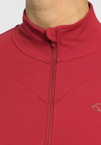 Rød lynlåsjakke med høj krave, blødt stof og syninger langs skuldrene. Har et lille logo på venstre side.