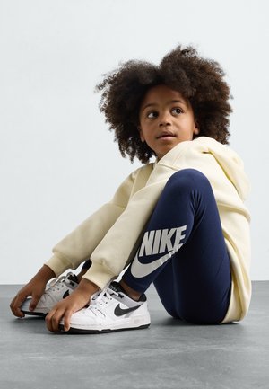 Világos sárga kapucnis pulóver, sötétkék leggings fehér Nike logóval, és fehér sportcipő fekete díszítéssel. Gyermek egy szürke felületen ül.