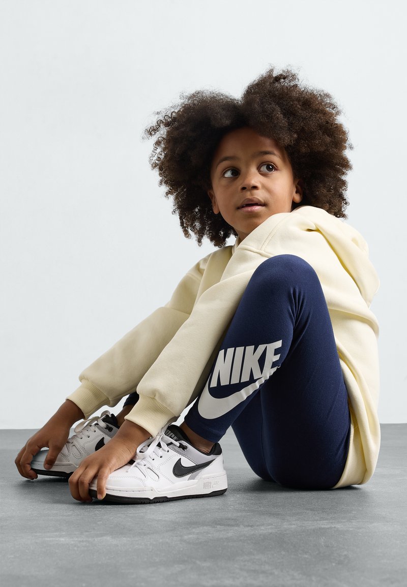 Svetlo rumena kapuca, mornarsko modre legice z belim logotipom Nike in bele superge z črnimi poudarki. Otrok sedi na sivi površini.