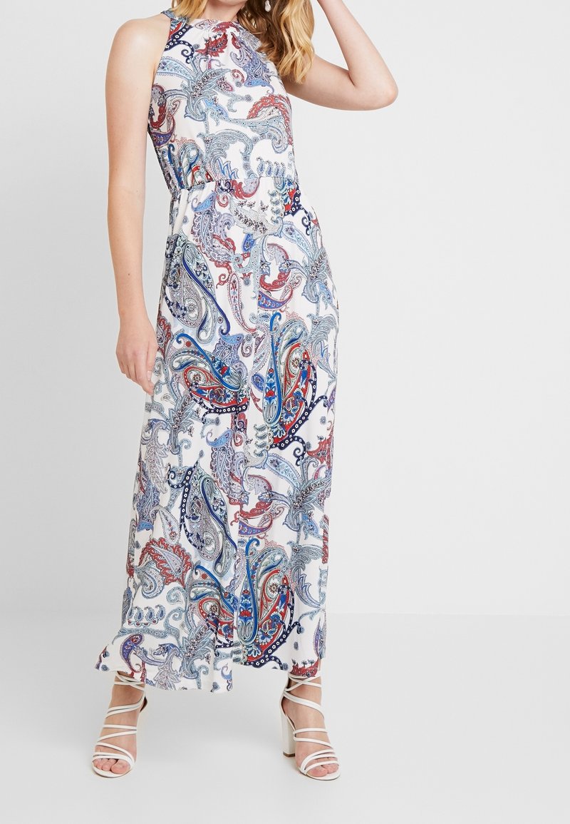 Femme portant une robe maxi blanche sans manches avec un imprimé cachemire bleu, rouge et gris, associée à des sandales blanches à talons hauts et à brides.