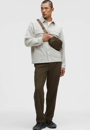 Moški, ki nosi svetlo sivo srajco z gumbi, olivno zelene hlače, črne superge in rjavo torbico čez ramo, stoji pred enostavnim ozadjem.