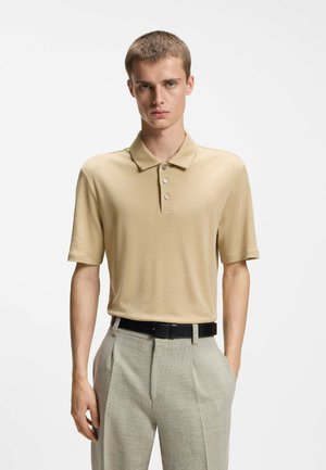 Jeune homme debout portant un polo beige à manches courtes rentré dans un pantalon gris clair avec une ceinture noire sur fond blanc.