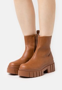 Stivali ankle in pelle marrone con suola piatta robusta. Presentano una zip laterale e un pull tab sul retro per facilitarne l'indossamento. Texture liscia.