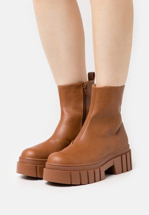 Botas de cuero marrón hasta el tobillo con una suela de plataforma gruesa. Tienen una cremallera lateral y una lengüeta en la parte posterior para un fácil uso. Textura suave.