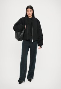Chaqueta bomber corta negra con cremallera plateada, combinada con jeans oscuros de pierna ancha. La modelo sostiene un bolso de cuero negro y está de pie frente a un fondo blanco.
