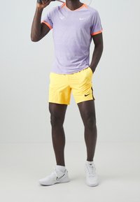 Lila träningsskjorta med orange detaljer, kombinerad med gula shorts som har en svart logotyp. Vita sneakers fulländar outfiten.