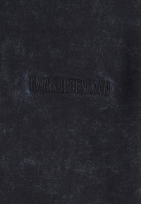 Tmavě modrý oděv s texturovaným povrchem, s jemnými variacemi odstínu. "HAN KJØBENHAVN" je vyražen černou nití.