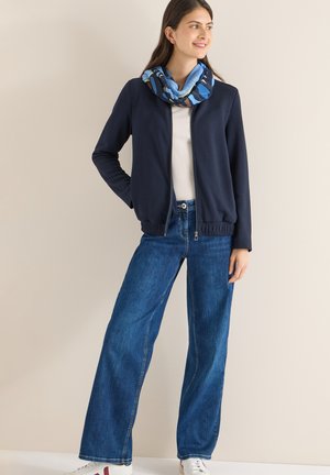 Donkerblauwe ritssluiting jas met elastische tailleband, wit overhemd eronder, wijde blauwe spijkerbroek en een meerkleurige, patroon sjaal.