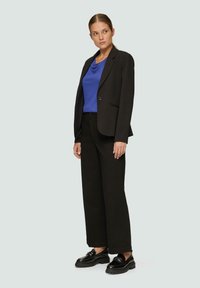 Zwarte blazer over een blauwe colletje-top, gecombineerd met zwarte wijde broek en zwarte chunky schoenen. Glad materiaal met gestructureerd ontwerp.