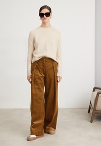 Beiger flauschiger Pullover, weite braune Cordhosen mit Falten und braunen Slippern. Große schwarze Sonnenbrille und Ohrstecker.