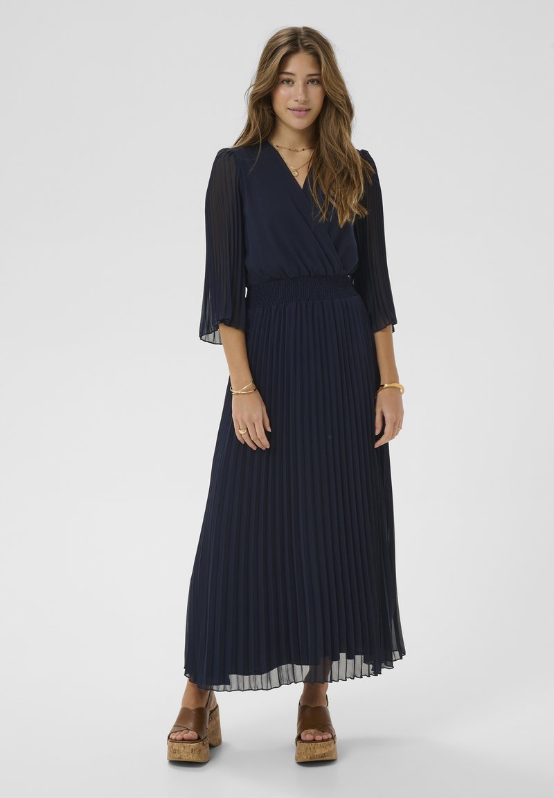 Marineblaues, plissiertes Maxikleid mit Wickeldesign und transparenten Glockenärmeln. Mit geraffter Taille und geschichtetem Stoff. Braune Plateausandalen.