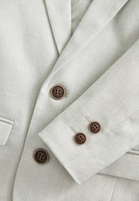 Blazer beige clair avec des boutons marron sur la manche et le devant, montrant la texture du tissu et les détails des boutons.