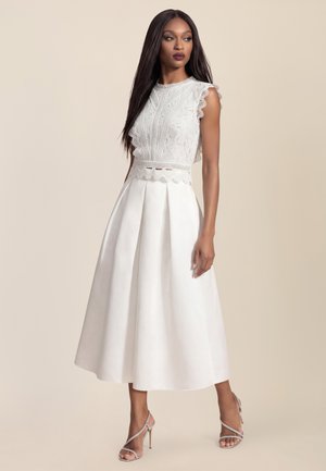 Frau trägt ein ärmelloses weißes Spitzentop und einen hoch taillierten plissierten weißen Midirock mit silbernen Sandalen mit hohem Absatz, stehend vor einem beigen Hintergrund.