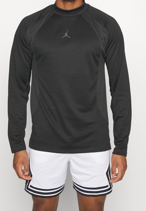 Man wearing black long-sleeve athletic top with subtle mesh panels and white basketball shorts with black stripes, standing against plain background.