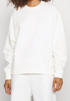 Person iført enkel hvid oversized sweatshirt og matchende hvide bukser, der viser overkrop og arme mod en ensfarvet baggrund.