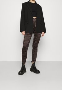 Blazer noir oversize, crop top noir ajusté, pantalon froncé marron brillant et bottines noires chunky, formant une tenue superposée et texturée.
