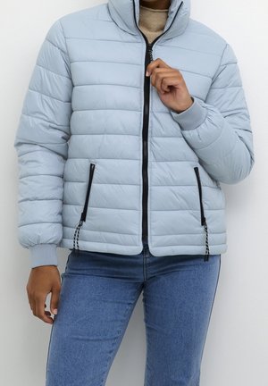 Persoon draagt een lichtblauwe doorgestikte jas met zwarte rits en zakken, gecombineerd met een blauwe spijkerbroek en een beige coltrui.