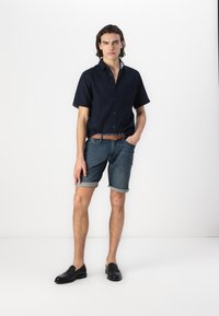 Chemise en denim bleu marine à manches courtes avec des boutons, associée à un short en denim à ourlet roulé, une ceinture en cuir marron et des mocassins noirs. Tenue simple et décontractée.