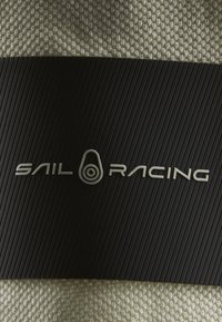 Černý a krémový textil s tkanou vrstvou nahoře a ribovaným černým pruhem. Pruh obsahuje embosovaný text "SAIL RACING" ve stříbrné barvě.