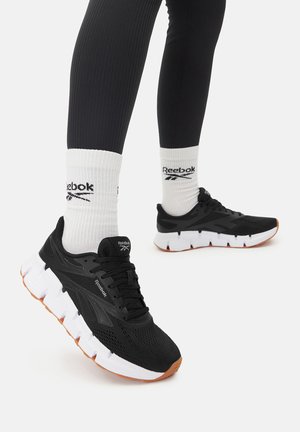 Czarne sportowe trampki z siateczkową cholewką, białą środkową podeszwą i pomarańczową gumową podeszwą zewnętrzną. Połączone z białymi skarpetkami z logo Reebok.