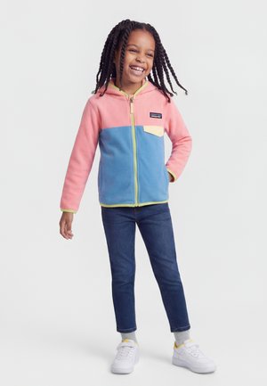 Χαμογελαστή κοπέλα με πλεγμένα μαλλιά στέκεται με το χέρι στη μέση, φορώντας ροζ και μπλε φλις ζακέτα με φερμουάρ, σκούρο τζιν και λευκά αθλητικά παπούτσια.