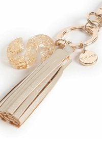 Llavero con una letra "S" de resina en tono dorado, un borlas de color beige claro hecha de material texturizado, y una etiqueta circular grabada con "MIRIADA."