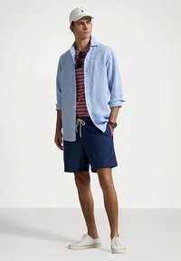 Šviesiai mėlyna palaidinė ant raudonos ir tamsiai mėlynos dryžuotos marškinėlių, tamsiai mėlyni šortai, balti sportbačiai ir balta kepurė. Kasdienis vasaros aprangos derinys.