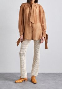 Blusa marrone testurizzata con nodo al collo, abbinata a pantaloni in crochet color crema e scarpe in pelle color sabbia. La blusa presenta una chiusura con bottoni e maniche ampie.