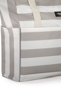 Beige és fehér csíkos anyagú táska elülső zsebbel és bézs vállpánttal, részleges nézet fehér háttér előtt.