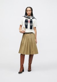 Γυναίκα που φοράει λευκό μπλουζάκι με κόκκινο κείμενο, μπεζ πλισέ φούστα, μπλε σκούρη και λευκή ριγέ ζακέτα δεμένη γύρω από τους ώμους, και καφέ μπότες έως το γόνατο.