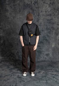Svart isolerad väst över en svart T-shirt, bruna kordjaktsbyxor och vita sneakers. Liten svart accessoarväska bärs crossbody. Brun keps.