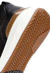 Zwarte sneakersole met een getextureerde tan rubberen buitenzool, voorzien van een witte tussenzool en glanzende bruine accenten. Zigzagprofiel zichtbaar.