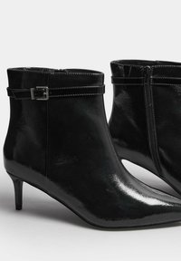 Botas de tobillo de charol negro con punta afilada, tacón pequeño, cremallera lateral y una correa con hebilla alrededor del tobillo. Textura suave.