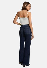 Pantalon en denim évasé de couleur bleu foncé avec une taille haute. Débardeur blanc à fines bretelles. Couture unique et deux poches arrière détaillées.