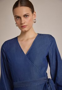 Denim omslagskjole i mørkeblå med lange ermer, V-hals og belte i livet. Glatt tekstur med minimale sømdetaljer.