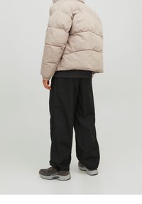 Vadderad, beige jacka med hög krage, elastiska ärmar och en kortad design, tillsammans med löst sittande svarta byxor och grå sneakers.