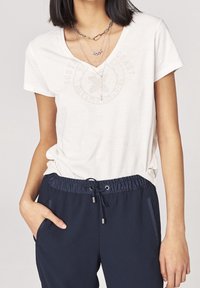 Camiseta blanca de manga corta con escote en V y un ligero detalle gráfico en el pecho, combinada con pantalones de cintura elástica azul marino con bolsillos.
