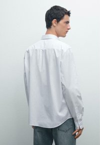 Camicia bianca a maniche lunghe con sottili strisce verticali, colletto con bottoni e orlo arrotondato. Indossata con jeans di denim blu chiaro.