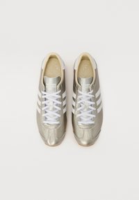 Ténis atléticos metálicos dourados com riscas brancas, fecho de atacadores, sola de borracha e parte superior texturizada. Design clássico e esquema de cores neutras.