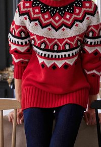 Pull rouge tricoté présentant un motif géométrique en crème et noir, avec un ourlet et des poignets côtelés, associé à un jean bleu foncé.