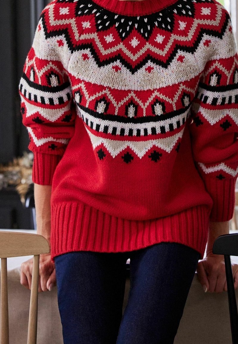 Pull rouge tricoté présentant un motif géométrique en crème et noir, avec un ourlet et des poignets côtelés, associé à un jean bleu foncé.