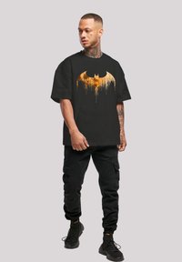 Melnā T-krekls ar lielu dzeltenu Betmena logotipu, kas ir ar pilienu efektiem. Kombinēts ar melniem cargo šortiem un tumšām apaviem.