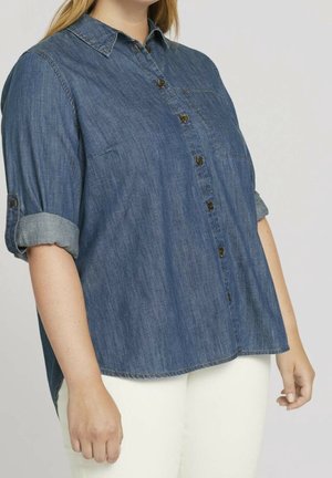 Vrouw draagt een blauw denim blouse met knopen en opgerolde mouwen en een witte broek, staand voor een effen achtergrond.