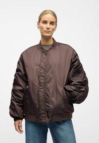 Brun bomberjakke med quilted design, ribbede ærmer og en lynlås lomme. Fremstillet af glat, let materiale med en løs pasform.